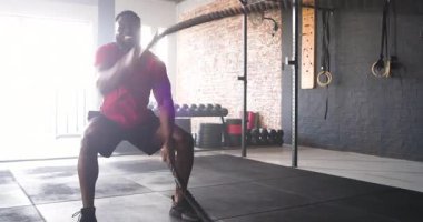 Afrika kökenli Amerikalı erkek, spor salonunda ipi sıkıca kavrıyor, sallıyor, sonra çömelmiş nefes alıyor. Güç, kondisyon, spor, spor, spor, spor, spor