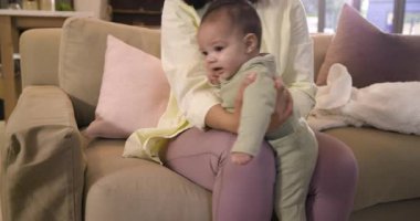 Anne kucağında küçük kızını evdeki kanepeye yatırıp dengeyi sağlamak için hoplayıp zıplatıyor. Bakıcı, yetiştirme, geliştirme, konforlu, aile içi, etkileşim, doğal ışık