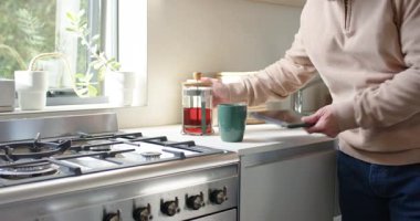 Olgun Asyalı bir adam mutfakta yeşil bardakta çay demliyor, Fransız pres makinesiyle, yudumluyor, tablet okuyor. Minimalist, dinginlik, sağlık, rahatlama, modern, yaşam tarzı, sağlık