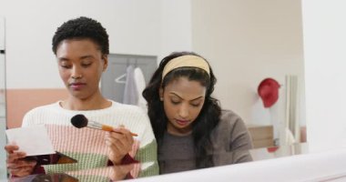 Kartla karıştırılmış Afrika kökenli Amerikalı kadın ve aynada kozmetik tüpü büken Hintli kadın. Güzellik, makyaj, kozmetik, cilt bakımı, kibir, zarafet, tarz