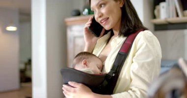 Evde telefonla konuşurken, bebek taşıyıcısında sallanan bir anne ve bir bebek var. Aile, annelik, bakıcılık, bakım, rahatlama, evcil, samimi