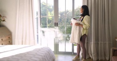 Beyaz battaniye tutuyor, kadın yatak odasına giriyor, kumaşı düzleştiriyor ve dışarı bakıyor, kopyalama alanı. Rahatlama, dinginlik, doğal ışık, konfor, tefekkür, iç mekan, minimalist