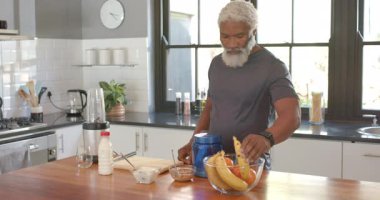 Kıdemli Afro-Amerikalı adam, mutfak adasında blender için muz soyarken ve parçalarken görülüyor. Sağlık, beslenme, smoothie, hazırlık, mutfak, yaşam tarzı, sağlık