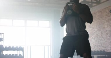 Afrika kökenli Amerikalı erkek sporcu, su ısıtıcısını teker teker kavrıyor ve pencerelerin önünde salona çömeliyor. Güç, zindelik, egzersiz, antrenman, spor, sağlık, spor