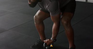 Afro-Amerikalı bir adam paspasa basıyor, kettlebell 'i rafa kaldırıyor sonra da salonun tepesine bastırıyor. Güç, antrenman, spor, spor, spor, atletizm, disiplin