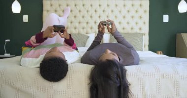 Afro-Amerikalı ve Hintli arkadaşlar yatakta uzanmış akıllı telefonları kaydırırken aynı zamanda gülüyorlar. Arkadaşlık, bağlantı, iletişim, rahatlama, rahatlama, yaşam tarzı, konforlu