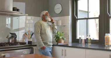 Olgun Afro-Amerikan adam mutfakta tezgaha yaslanıyor, akıllı telefon görüşmeleri yapıyor, haberlere gülümsüyor. Gündelik, modern, canlı, yaşam tarzı, yerli, rahat, iletişim