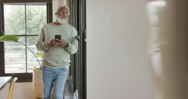 Son sınıf Afro-Amerikan erkeği oturma odasında pencereye yaslanmış düşünüyor ve akıllı telefonları kaydırıyor. Son sınıf, eğlence, teknoloji, yerli, rahat, çağdaş, yaşam tarzı