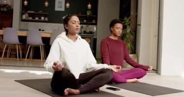 Değişik kadınlar sakinleşmek için meditasyon yapıyor gözlerini kapatıyor evdeki bitkilerin yanında yoga minderlerine el kaldırıyor. Meditasyon, farkındalık, dinginlik, rahatlama, sağIık, sükunet, ev