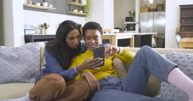 Afro-Amerikan ve Hintli kadınlar kanepeye yaslanmış akıllı telefonları kaldırıyor, fotoğrafları keşfediyorlar. Dost canlısı, dijital, güncel, çağdaş, kadınsı, bağlantı, rahatlamış