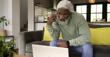 Afro-Amerikalı son sınıf öğrencisi akıllı telefonu dengeliyor sonra da oturma odasındaki çağrı sırasında dizüstü bilgisayarda yazıyor. Profesyonel, teknoloji, modern, gündelik, ev, yaşam tarzı, konsantre