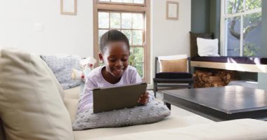 Afro-Amerikalı kız oturma odasındaki kanepeye yerleşirken tablet açısını ayarlıyor ve tıkırdatıyor. Teknoloji, çocukluk, rahatlama, iç mekan, aile, dijital, samimi