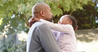 Afrikalı Amerikalı baba, koşan kızı kaldırıyor öpüşüyor ve arka bahçedeki ağacın altında kıkırdıyor. Aile, bağ, sevgi, şakacı, açık hava, doğa, mutluluk