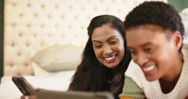 Çeşitli bayan arkadaşlar tablet direğini, kaydırma cihazlarını görüyor, gülüyor ve yatağı işaret ediyorlar. Arkadaşlık, teknoloji, rahatlama, iletişim, konfor, gündelik, bağlantı