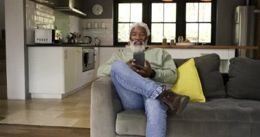 Kanepede oturan Afro-Amerikalı adam akıllı telefonu kaldırıyor, içeriği kayıyor, mutfağa bakıyor. Günlük, rahatlama, dijital, modern, yerli, yaşam tarzı, konfor
