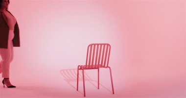 Duraklayan, spor ceketli bir kadın pembe stüdyodaki kırmızı sandalyeye yürüyor, rahat poz vermek için sırtüstü uzanıyor. Modern, moda, minimalist, zarafet, güven, stüdyo, portre