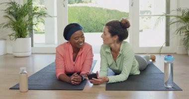 Çeşitli bayan arkadaşlar spor salonundaki paspaslarda egzersiz yapıyor telefon ekranlarını paylaşıyor ve biberonlarını yudumluyorlar. Dostça, sağlıklı, yaşam tarzı, rahatlama, işbirliği, doğal, modern