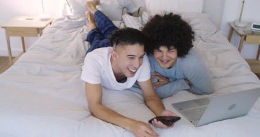 İki yetişkin erkek yatak odasında uzanıyor, akıllı telefon içeriğini paylaşıyorlar, dizüstü bilgisayarda yazıyorlar, sarılıyorlar. Samimiyet, bağlantı, rahatlama, modern, samimi, şefkat, arkadaşlık