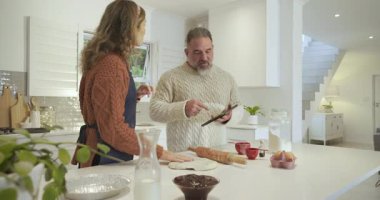 Mutfağa giren bir çift, tablet kullanan bir adam ve yemek pişirmek için hamur şekillendiren bir kadın. Aile içi, işbirliği, aşçılık, teknoloji, yaşam tarzı, konforlu, aile