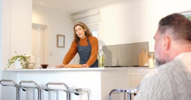 Yetişkin bir kadın, mutfak adasındaki yemek tariflerini nazikçe gülümseyerek anlatıyor. Aile içi, aşçılık, yaşam tarzı, modern, samimi, sıcak, davetkar.