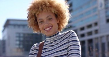 Çizgili süveter giymiş Afro-Amerikalı bir kadın, gülümsüyor ve rüzgar saçlarını dalgalandırırken şehir meydanında eğiliyor. Portre, özgüven, moda, zarafet, gündelik, yaşam tarzı, mimari