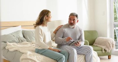 Çift yatak odasında oturuyor, adam tablet tıkırdatıyor, fotoğraf çekiyor, kadın el kol hareketi yapıyor ve gülüyor. Romantizm, samimiyet, rahatlama, yaşam tarzı, konfor, iletişim, bağlantı