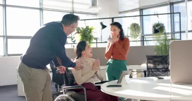 Çeşitli iş arkadaşları tableti inceliyor ve modern ofiste heyecan verici haberleri tekerlekli sandalyeyle kutluyorlar. İşbirliği, çeşitlilik, başarı, takım çalışması, yenilik, erişilebilirlik, pozitif
