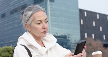 Kafe masasındaki akıllı telefonu dinleyen kıdemli bir kadın haberleri kontrol ediyor ve kahve fincanına ulaşıyor. Kentsel, yaşam tarzı, samimi, modern, teknoloji, rahatlama, tarz