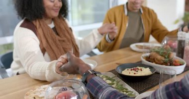 Afro-Amerikalı çift yemek masasında ellerini tokuşturup başlarını eğerek şükranlarını sunuyorlar. Minnettarlık, birliktelik, kutlama, sıcaklık, huzur, aile, gelenek.