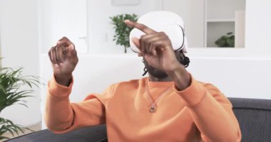 Adam kanepeye oturmuş, beyaz VR kulaklık takıyor, sanal panelleri çimdikliyor, sonra da duruşu ayarlıyor. Teknoloji, yenilik, sürükleyici, fütüristik, eğlence, dijital, etkileşim