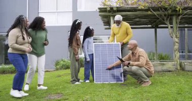 Erkek teknisyenler tablet tıkırdatıyor ve çeşitli ailelere rehberlik ediyor. Ön bahçede güneş paneli kablosunu paylaşıyorlar. Güneş enerjisi, yenilenebilir, sürdürülebilirlik, açık hava, işbirliği, topluluk, modern tasarım