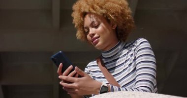 Telefondan mesaj geliyor, Afro-Amerikalı bir kadın beton gölgeliğin altında oturmuş gülüyor. Kentsel, gelişigüzel, modern, teknoloji, iletişim, rahatlama, samimiyet