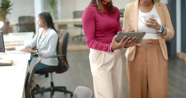 Çeşitli iş arkadaşları kahvelerini yudumlarken ve ofiste yazı yazarken tabletin içeriğine dokunuyorlar. İşbirliği, takım çalışması, iletişim, yenilik, modern, profesyonel, üretkenlik