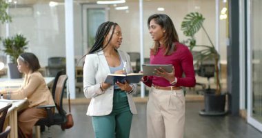 Çeşitli kadın iş arkadaşları ofis koridorunda yürüyor, tablet ekranı işaret ediyor ve not defterine yazıyor. İşbirliği, takım çalışması, iletişim, yenilik, modernlik, profesyonel, üretkenlik