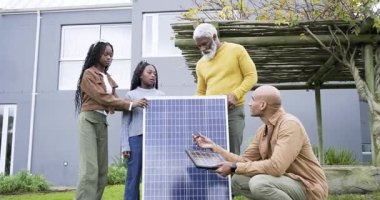 Teknisyen avluda diz çöküp güneş paneli kablolarını panele bağlarken Afro-Amerikan aile izliyor. Sürdürülebilirlik, yenilenebilirlik, çevre dostu, eğitim, yenilik, modern, açık hava