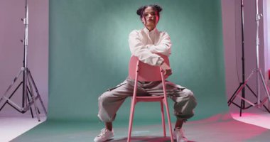 Stüdyoda genç bir kadın, rahat bir şekilde sandalyede oturuyor, sokak kıyafetleri giyiyor. Moda, tarz, güven, moda, giysi, şehir