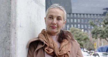 Şehir kaldırımında duvara yaslanmış, kahverengi ceketli, eşarplı, gülümseyen olgun beyaz bir kadın. Portre, kentsel, günlük, moda, sokak, yaşam tarzı, pozitiflik