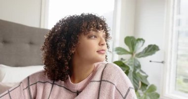 Yatakta oturan, güneş ışığını hisseden ve kameraya gülümseyen bir kadın. Rahat, huzur, doğal ışık, iç mekan, düşünceli, minimalist, kadınsı