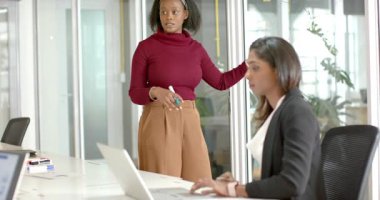 Ofis toplantısında siyahi kadın cam tahtayı gösterirken Hintli meslektaşımız dizüstü bilgisayarda yazı yazıyor. İşbirliği, takım çalışması, yenilik, şirket, profesyonel, beyin fırtınası, modern