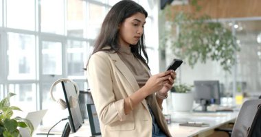 Ofis masasında iş kıyafeti giymiş Hintli bir kadın, telefonda mesajları kaydırıyor, güncellemede gülümsüyor. Profesyonel, modern, minimalist, işyeri, teknoloji, iletişim, yaşam tarzı