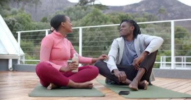 Afro-Amerikalı çiftler gülümseyerek ahşap teras yoga minderlerinde su içmek için su paylaşıyorlar. Sağlık, rahatlama, bağlantı, açık hava, arkadaşlık, uyum, huzur.