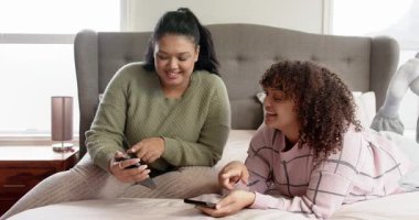 Anne ve kızı telefonlarını dinliyor, videoları gösteriyor, telefonları alıyor ekranları değiştiriyor ve yatakta gülüyorlar. Aile, bağ, rahatlama, teknoloji, samimiyet, samimiyet, samimiyet.