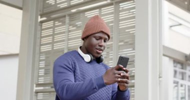 Afro-Amerikalı adam otobüs durağına örgü şapkayla giriyor, kulaklık takıyor, akıllı telefon kaldırıyor, gelişini kontrol ediyor. Kentsel, teknoloji, gündelik, modern, sokak giyimi, iletişim, yaşam tarzı