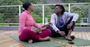 Balkon minderlerinde oturan Afro-Amerikalı çift göz teması kuruyor. Omuz omuza veren adam gülüyor. Sağlık, bağlantı, rahatlama, açık hava, arkadaşlık, farkındalık, mutluluk.