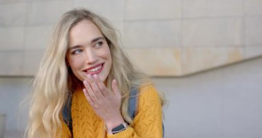 Kamera dışından sesler duyan, gülen beyaz kadın taş duvarda saçlarını ören, sırt çantası görünen. Neşe, samimiyet, yaşam tarzı, açık hava, günlük, ifadeler, sükunet