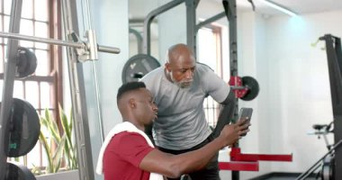 Afrika kökenli Amerikalı antrenör, halter rafında kayan müşterileri telefonla işaret ediyor. Fitness, koçluk, motivasyon, güç, takım çalışması, kararlılık, refah