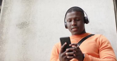 Akıllı telefonu dinleyen, turuncu kazaklı bir adam gri duvarda kulaklıkla müzik çalıyor. Teknoloji, kentsel, gündelik, rahatlama, açık hava, yaşam tarzı, iletişim