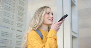 Süveter ve sırt çantası giyen, telefondan ağıza konuşan ve apartmanda asistanla konuşan bir kadın. Gelecekçi, kentsel, gösterişli, iletişim, teknoloji, yaşam tarzı, çağdaş