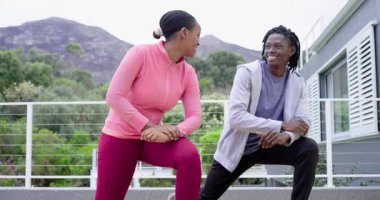 Göz temasını paylaşan Afrikalı Amerikalı arkadaşlar terasta hareketler ve gerinmelerle ısınıyorlar. Açık hava fitness, esneme, yoldaşlık, atletik, modern mimari, refah, aktif yaşam tarzı