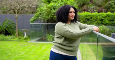 Afrika kökenli Amerikalı bir kadın balkona çıkıyor korkuluklara yaslanıyor bahçeye bakıyor ve gülüyor. Dinginlik, rahatlık, düşünce, samimiyet, doğal, açık hava, yaşam tarzı.